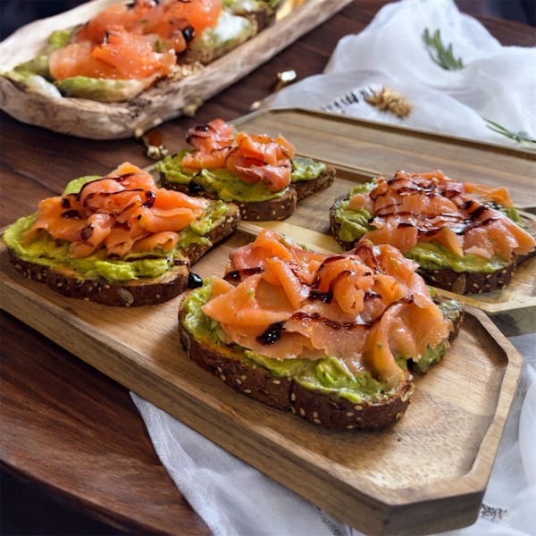 Four avocado toasts topped with smoked salmon and balsamic glaze on wooden trays.