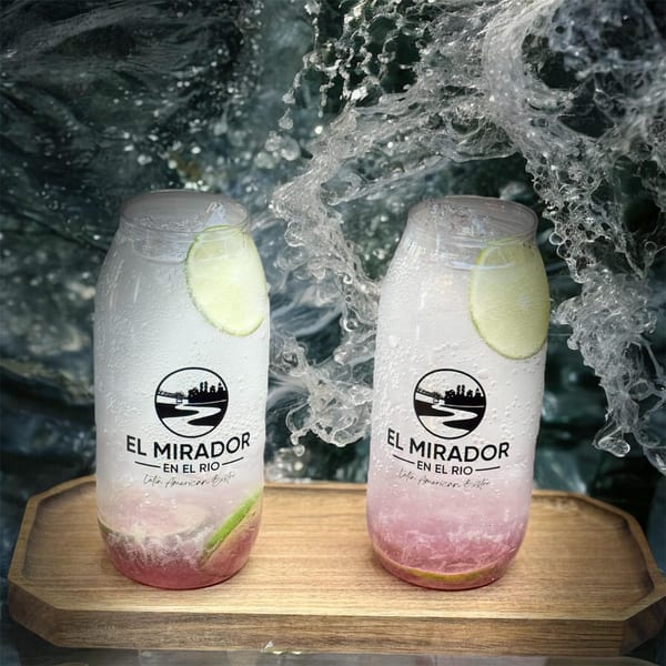 Two El Mirador En El Rio drinks with lime slices and pink liquid on a wooden tray, with splashing water.