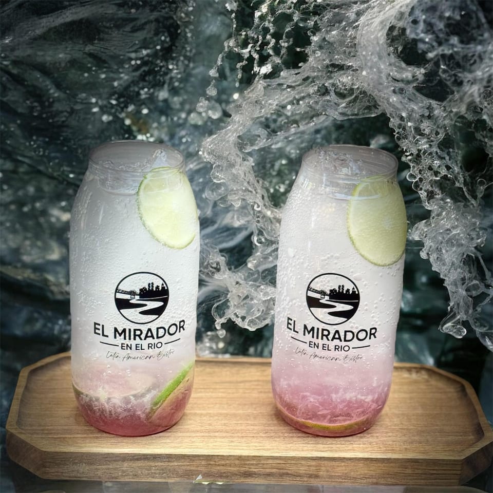 Two El Mirador En El Rio drinks with lime slices and pink liquid on a wooden tray, with splashing water.