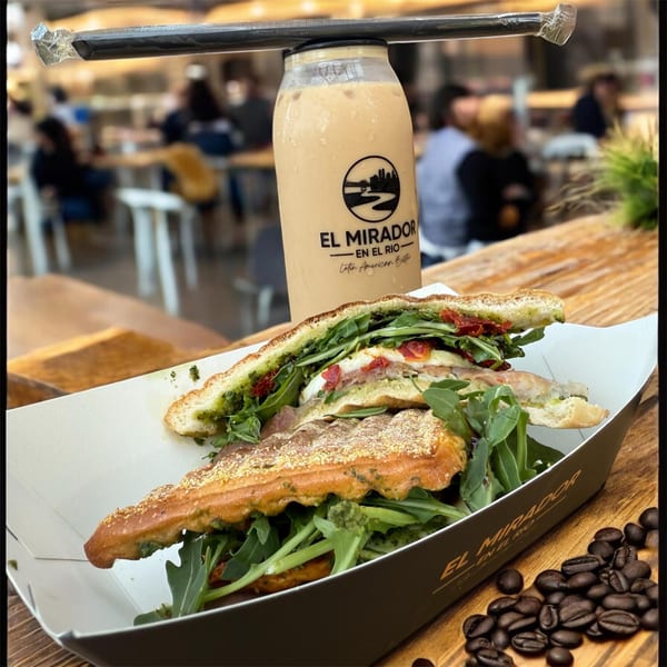 A sandwich with pesto, arugula, and sun-dried tomatoes, next to an El Mirador iced coffee.