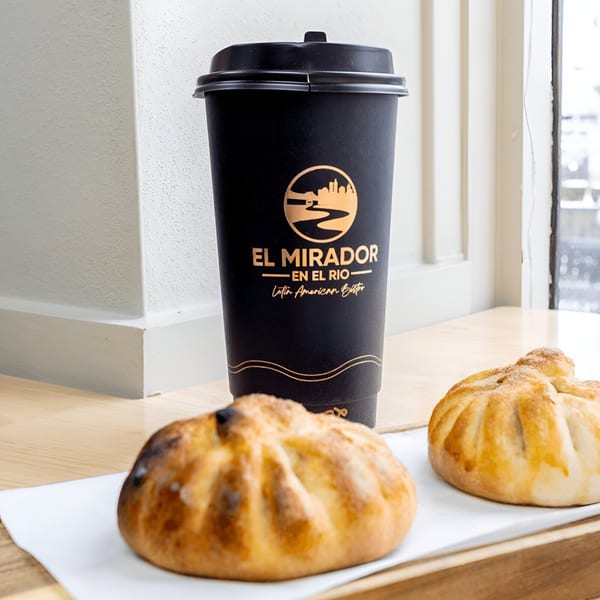 Two golden-brown Latin American pastries and a black El Mirador En El Rio coffee cup on a wooden tray.